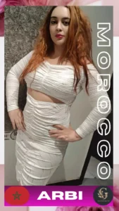 Moroccan woman in a stylish white ruched cut-out dress posing confidently indoors against a textured wall, with long wavy reddish-brown hair. Text on image says "MOROCCO" vertically and "ARBI" with a Moroccan flag and Asian Golden Spa logo. She is a professional Arbi-speaking therapist at Asian Golden Spa, Ras Al Khaimah.
