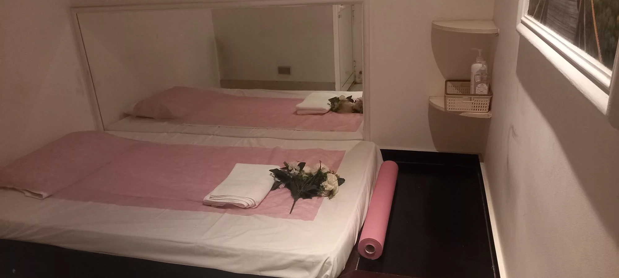 Massage room at Asian Golden Spa, Nakheel Prime Hotel Ras Al Khaimah, featuring a single bed with pink and white sheets, fresh flowers, towel, wall mirror, pink disposable bed sheet roll, shelf with sanitizer, and a nature-themed wall photo.