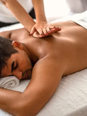 A shirtless man lying face down on a white massage bed with a towel under his head, receiving a deep tissue back massage from a female therapist applying pressure with both hands on his upper back.
