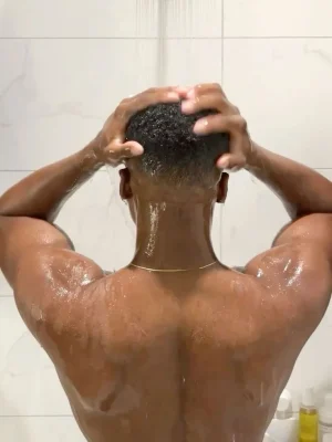 Back view of a muscular man taking a refreshing shower, hands massaging his head under a rainfall shower in a modern white-tiled bathroom, highlighting relaxation and hygiene — Asian Golden Spa, Ras Al Khaimah, Nakheel Prime Hotel, 1st Floor. Contact: 056 978 9770.