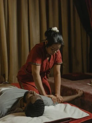 "Female Thai massage therapist in red traditional uniform performing arm and shoulder pressure technique on a male client lying face-up, at Asian Golden Spa, Nakheel Prime Hotel, Ras Al Khaimah"