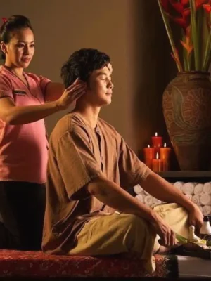 "Female massage therapist in pink uniform performing traditional Thai head massage on seated male client wearing brown spa clothing at Asian Golden Spa, Ras Al Khaimah, Nakheel Prime Hotel"